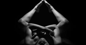 Black and white photograph showcasing an artistic nude pose with dramatic lighting and shadows.